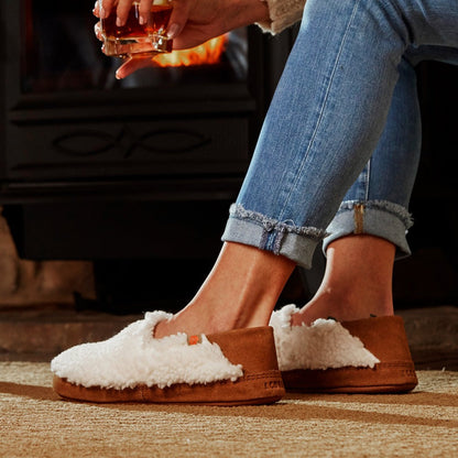 Women’s Acorn Moc with Collapsible Heel Slipper in Buff Popcorn on model sitting by a fireplace with a drink in her hand#color_buff-popcorn